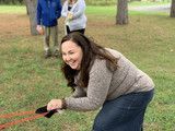 Woman in gray sweater crouches on grass, smiling while holding a red toy or leash in a park.