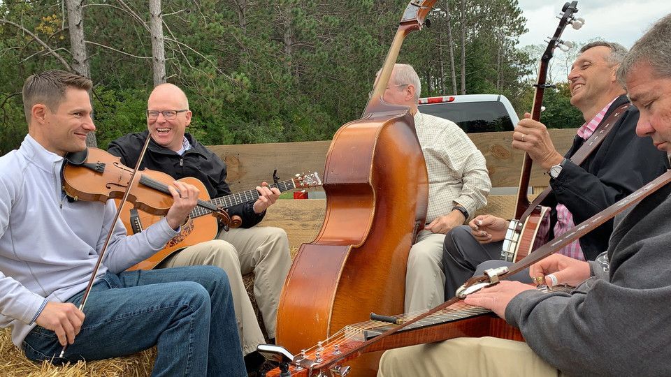 Musicians seated outdoors playing string instruments around a large upright bass on a road.