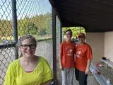 Three people standing by a chain-link fence, two wearing orange shirts and one in a neon yellow shirt, smiling outside.
