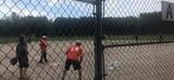 People standing on a field near a chain-link fence and goal, with one in a red shirt facing the others.