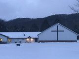 Snow-covered church buildings at dusk, with lit windows and a dark treeline in the background