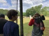 People outdoors taking photos near a field, with one holding a camera up to their face.