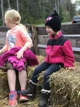 Two children sitting on hay bales outdoors, one in a pink jacket and one in a black-and-pink jacket
