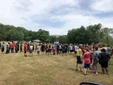 Large crowd gathered on a grassy field with trees in the background, some people standing and watching.
