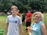 Two smiling people with water buckets stand outdoors in a park, with more people and greenery behind them