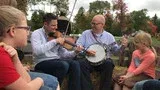 People sitting outdoors, playing banjo and guitar in a casual circle at a picnic table