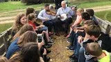 Group sitting outdoors in a circle, listening to a man speaking in the center.