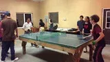 People playing table tennis in a room with a green ping-pong table and several spectators nearby