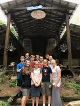 Group of people posing under a rustic wooden shelter with a sign overhead, outdoors in a forested area.
