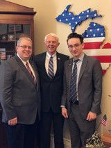 Three men in suits posing indoors beside a blue Michigan-shaped wall decoration and flags.