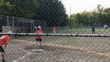 Baseball players on a dirt field behind a chain-link fence, with one batter in a red jersey at home plate.