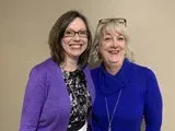 Two smiling women standing close together, one in a purple sweater and one in blue, against a plain wall