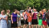 Group of people outdoors cheering and raising their arms, with trees in the background.