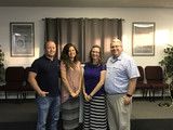 Four people posing together in a meeting room with chairs and framed wall art behind them.