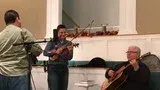 Three musicians performing in a room, with a violinist at center and a seated guitarist at right.