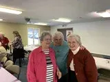 Three smiling people standing together in a bright indoor room with tables and other guests in the background