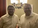 Two men in light shirts smiling indoors beneath a chandelier