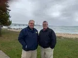 Two people standing on a grassy shore with water and boats in the background