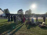People gathered outdoors at sunset near a building, with long shadows on the grass.