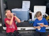 Two children standing by a truck, one in red drinking from a cup and one in blue looking down at a phone