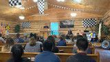 Conference hall with seated audience facing a speaker at a podium under warm wood-paneled walls.