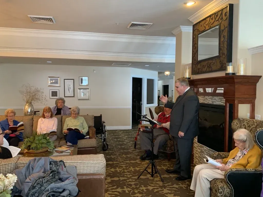 Man speaking to seated audience in a living room with a fireplace and large mirror