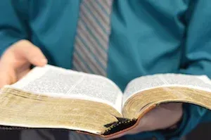 Hands holding an open book with yellowed pages against a teal shirt and striped tie