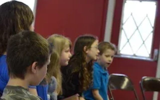Children seated in a classroom, facing forward near a red wall and window