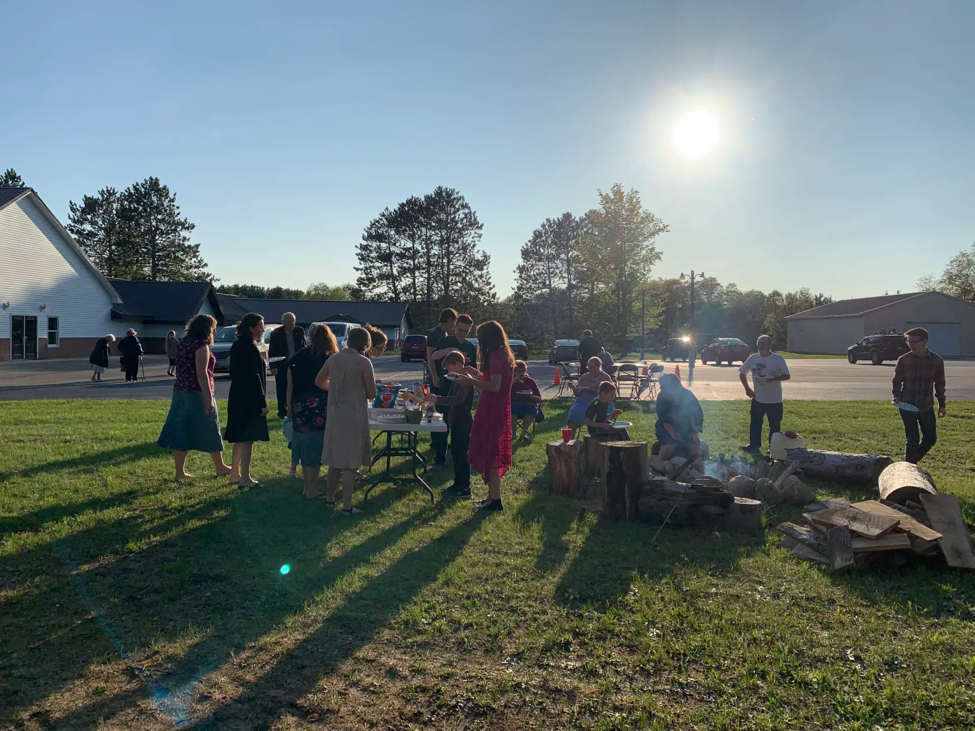 People gathered outdoors on a sunny lawn near a white tent, with long shadows and trees in the background.