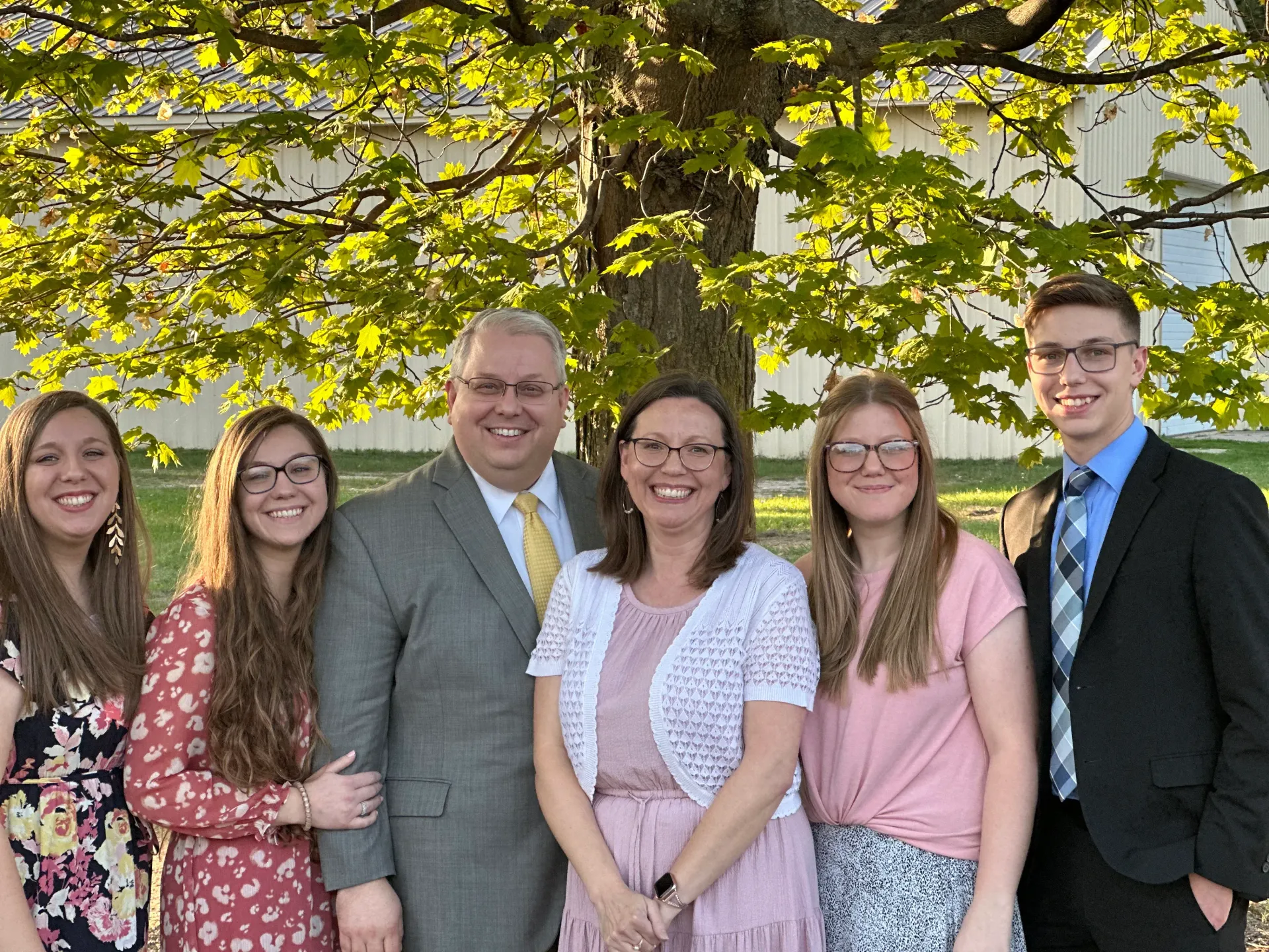 Six people smiling outdoors under a tree with yellow leaves, dressed in semi-formal attire.