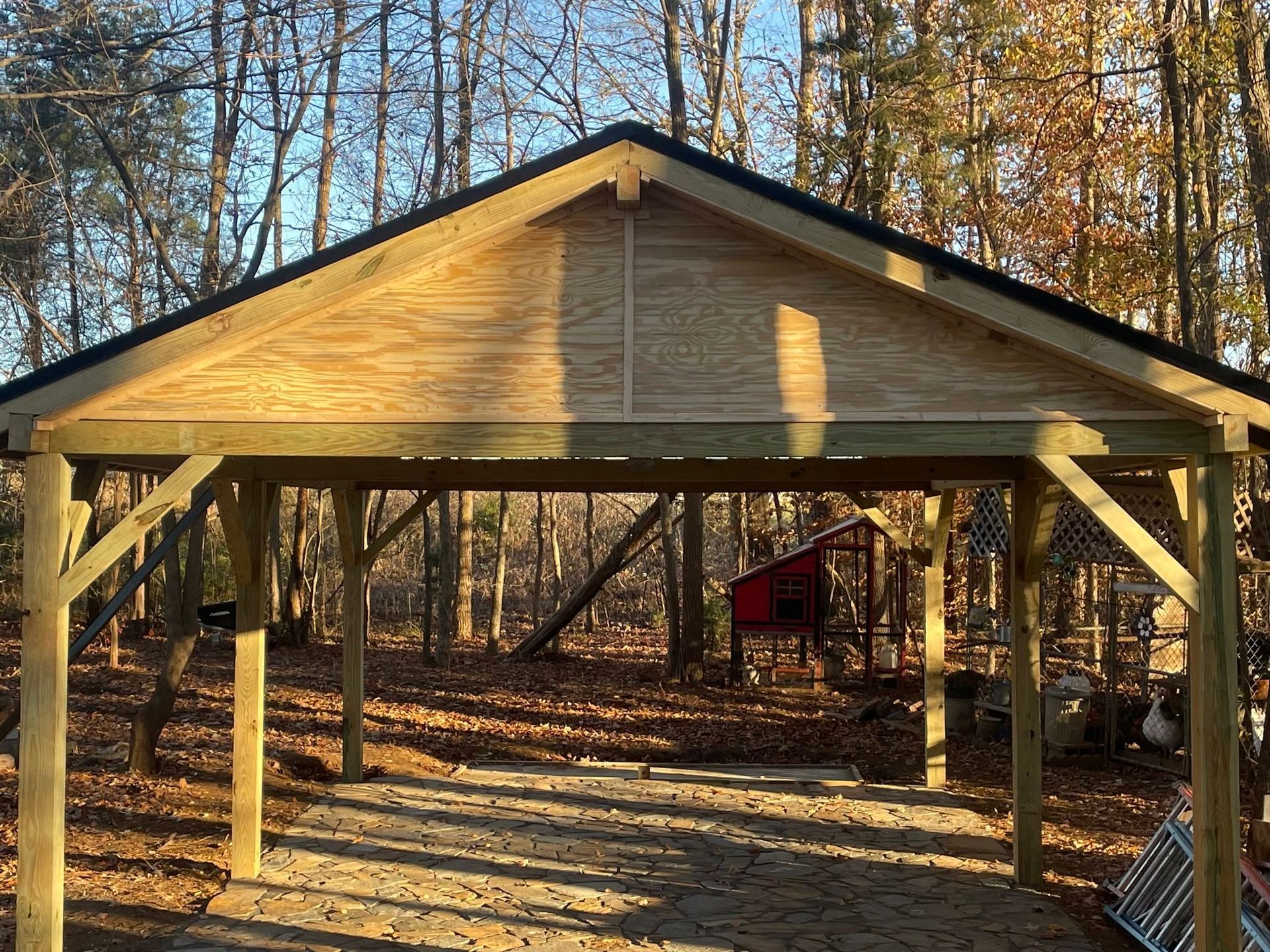 Wooden pavilion with a gabled roof in a wooded yard, lit by low sunlight.