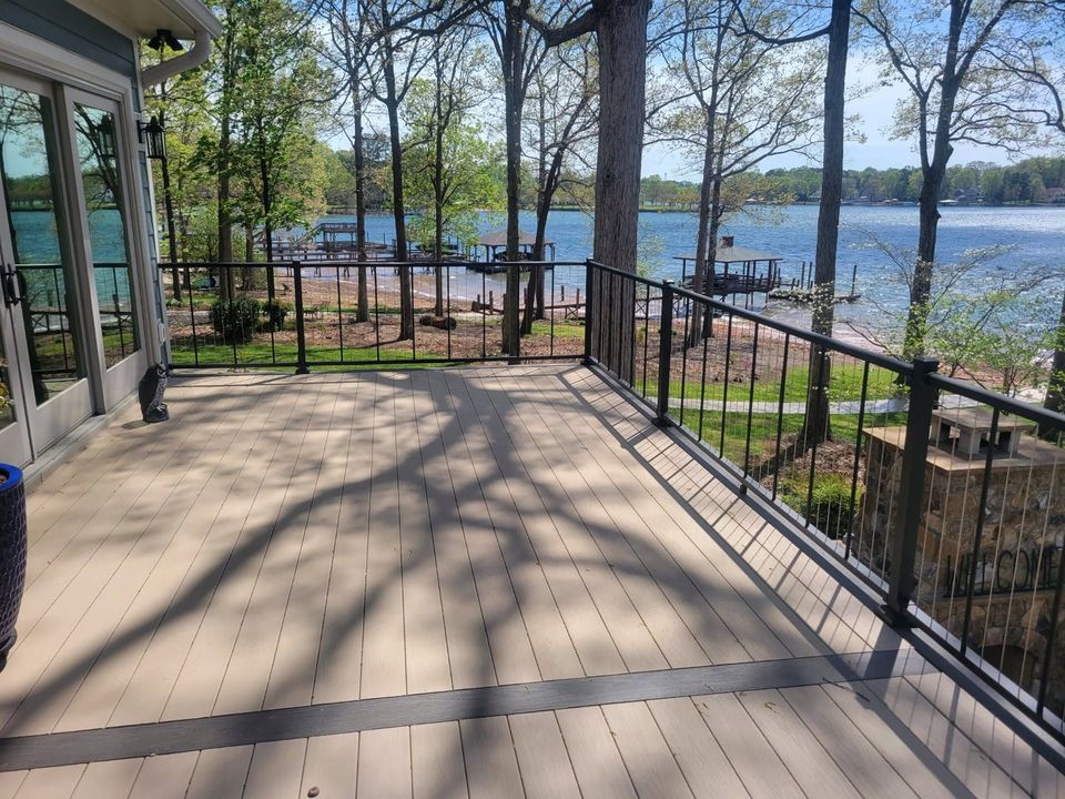 Wooden deck with black railing overlooking a lake, trees, and a sandy beach on a sunny day