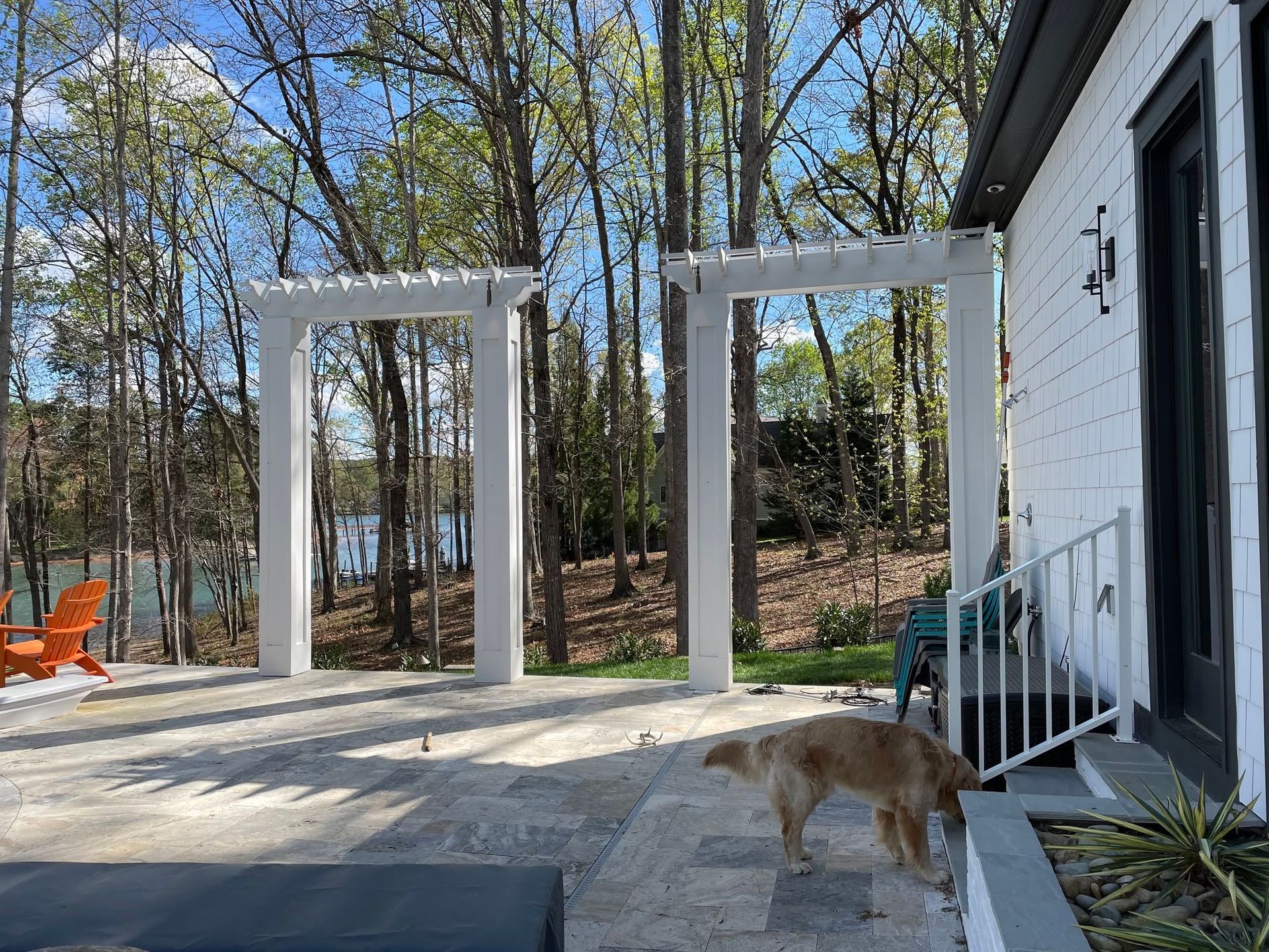 Patio with white pergolas, stone pavers, trees, and a tan dog near a house door