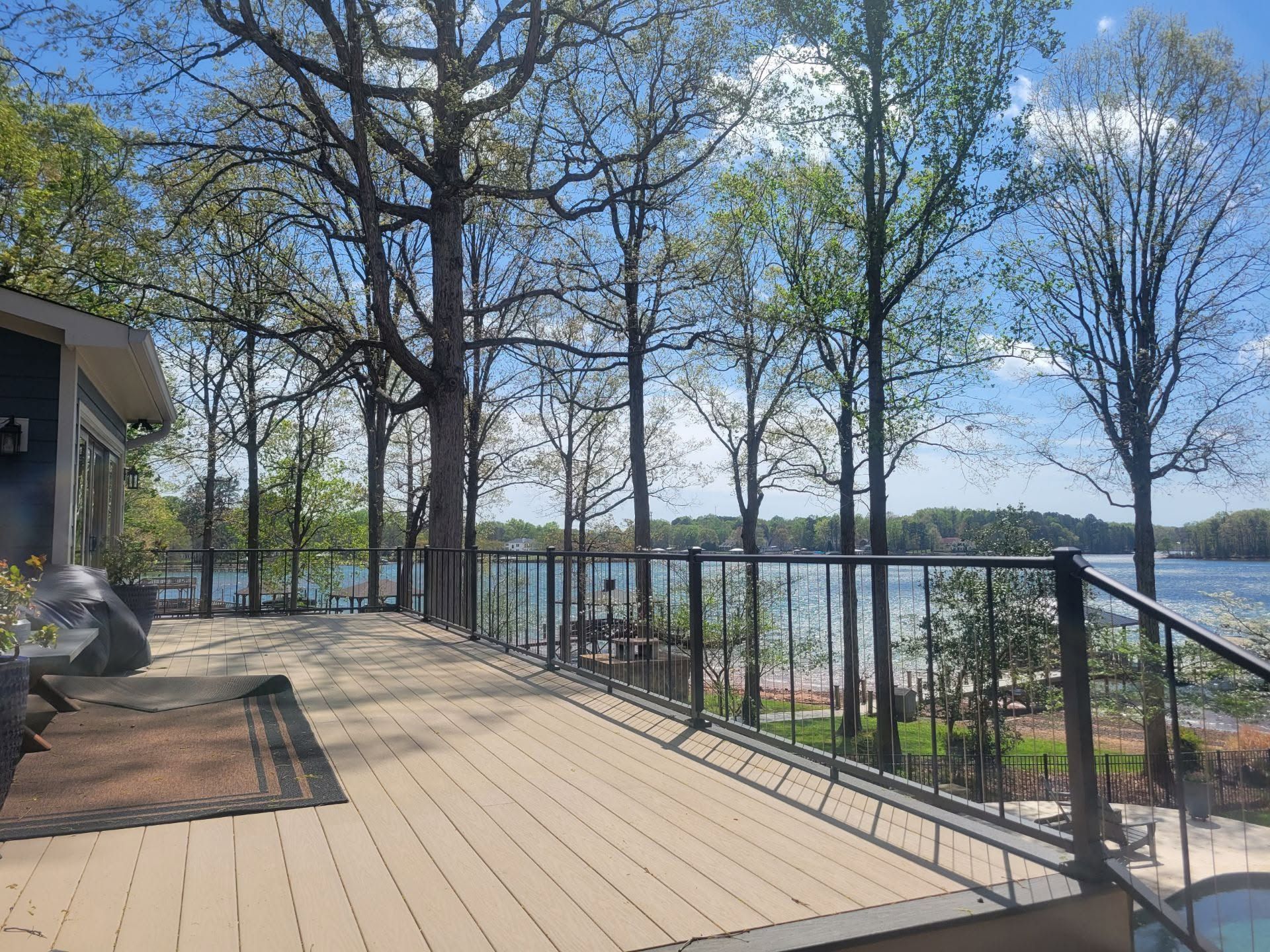 Wooden deck with black railing overlooking a lake, framed by leafless trees under a blue sky.