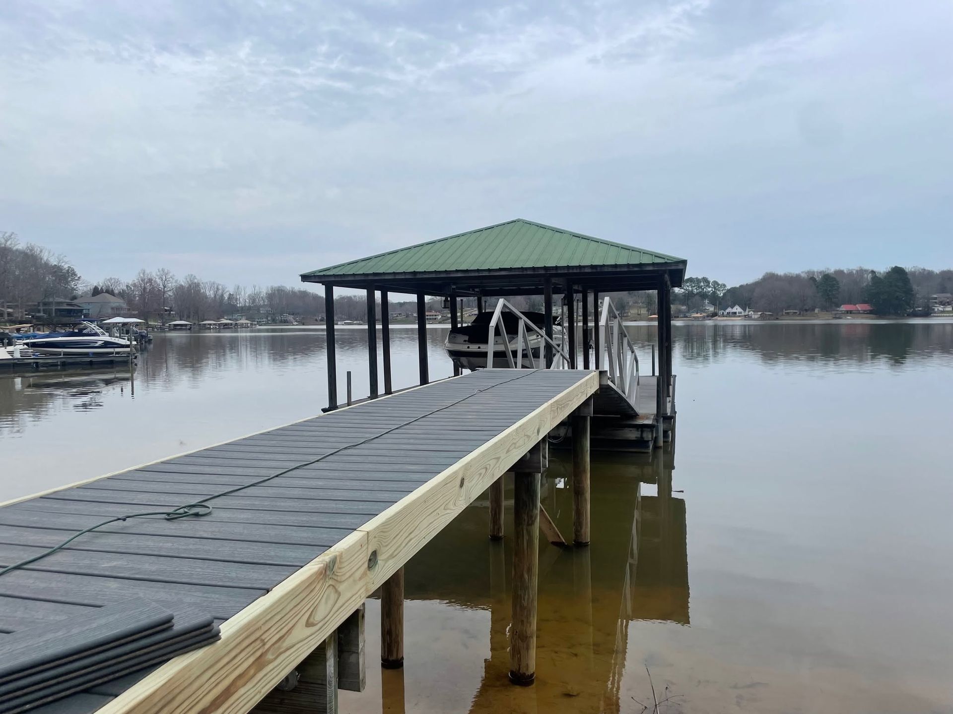 Wooden dock extending to a green-roofed boathouse on a calm lake under an overcast sky