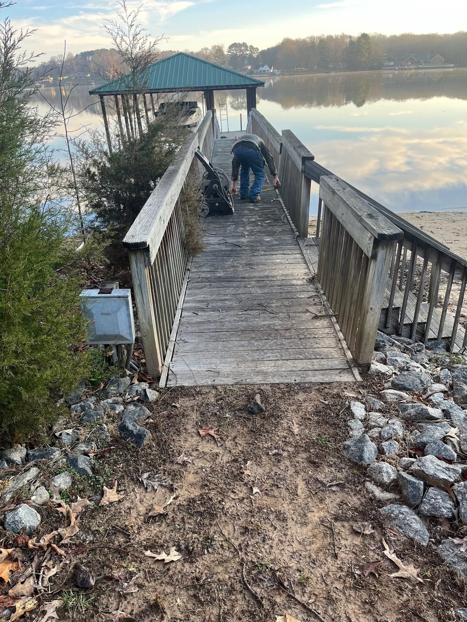 Wooden lakeside pier with a green-roofed gazebo, two people, and calm water at sunrise.