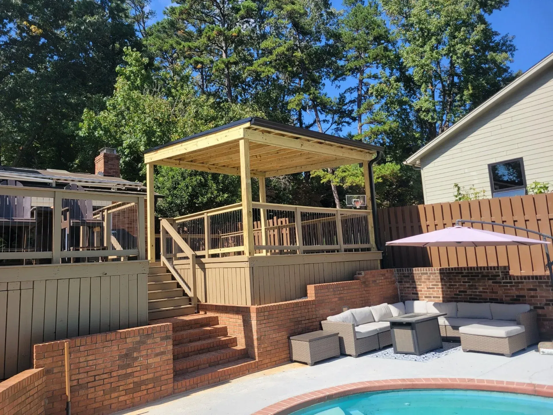 Backyard deck with pergola, wooden railing, brick steps, and gray patio seating under trees