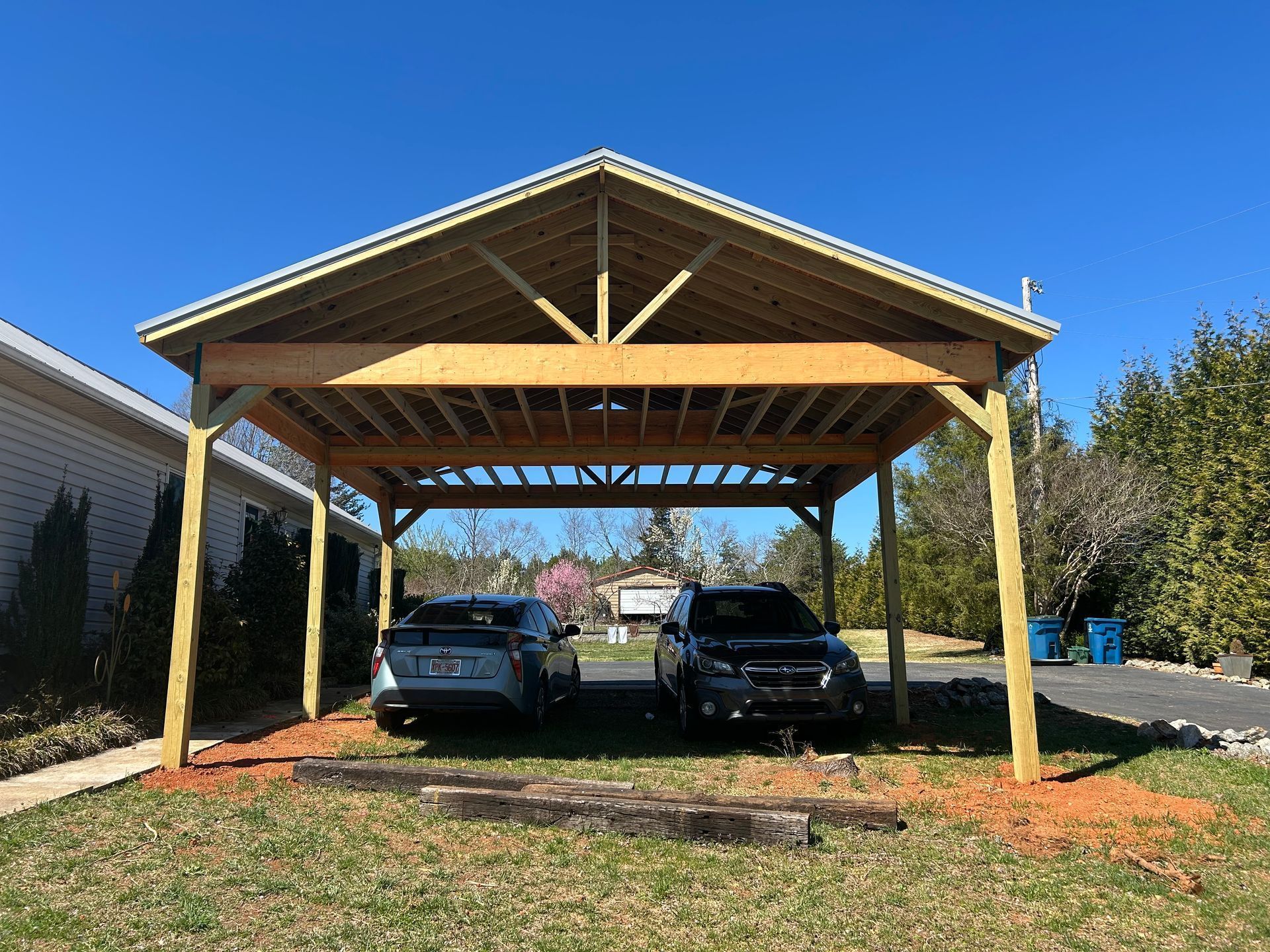 Two cars parked under a wooden carport in a sunny yard with green grass and trees.