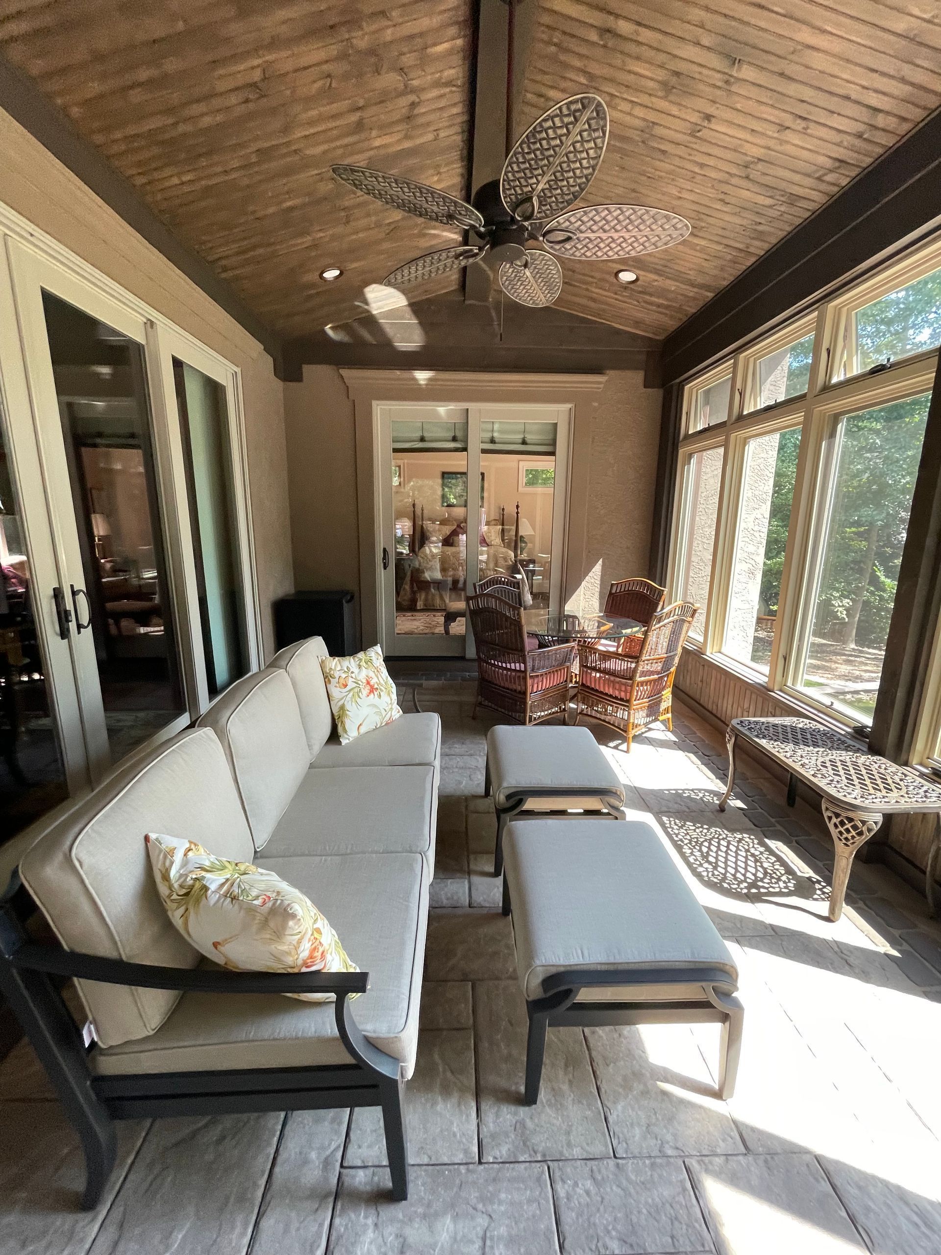 Sunroom with white sofa, gray ottoman, wicker chairs, and ceiling fan, lit by large windows