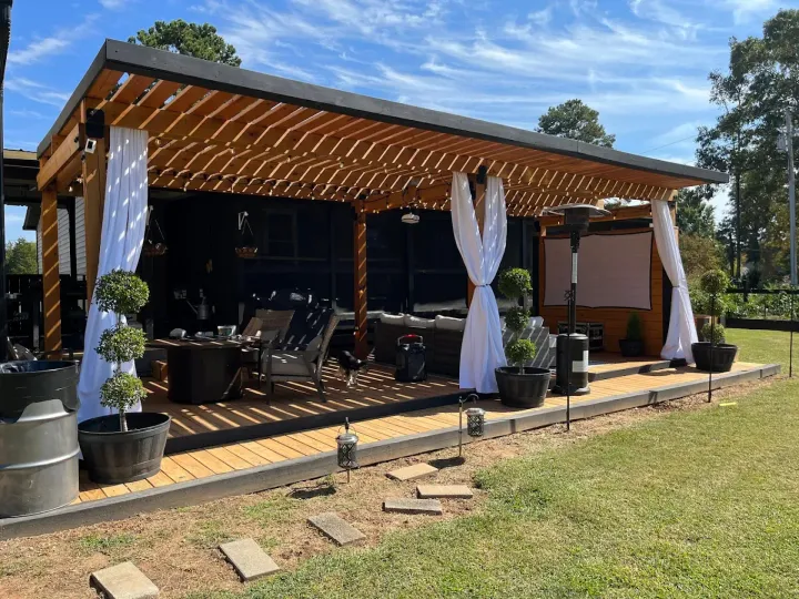 Outdoor patio with wooden pergola, sheer white curtains, seating, and potted plants on a sunny day