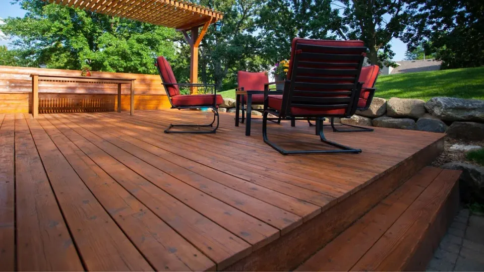 Wooden deck with red patio chairs under a pergola, beside a grassy yard and stone wall.