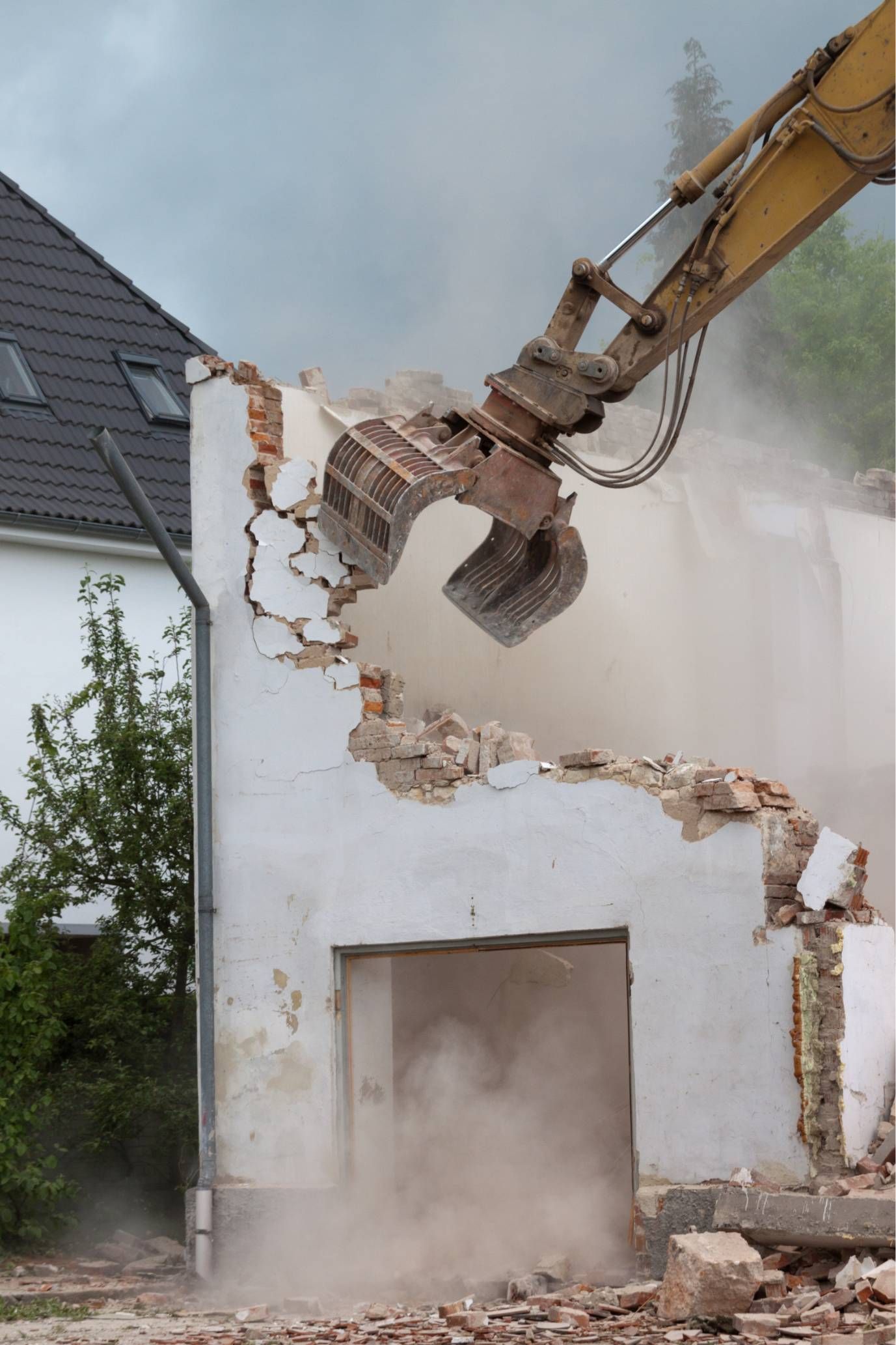 Excavator demolishing a white house wall, with dust and debris falling beside a sloped-roof home