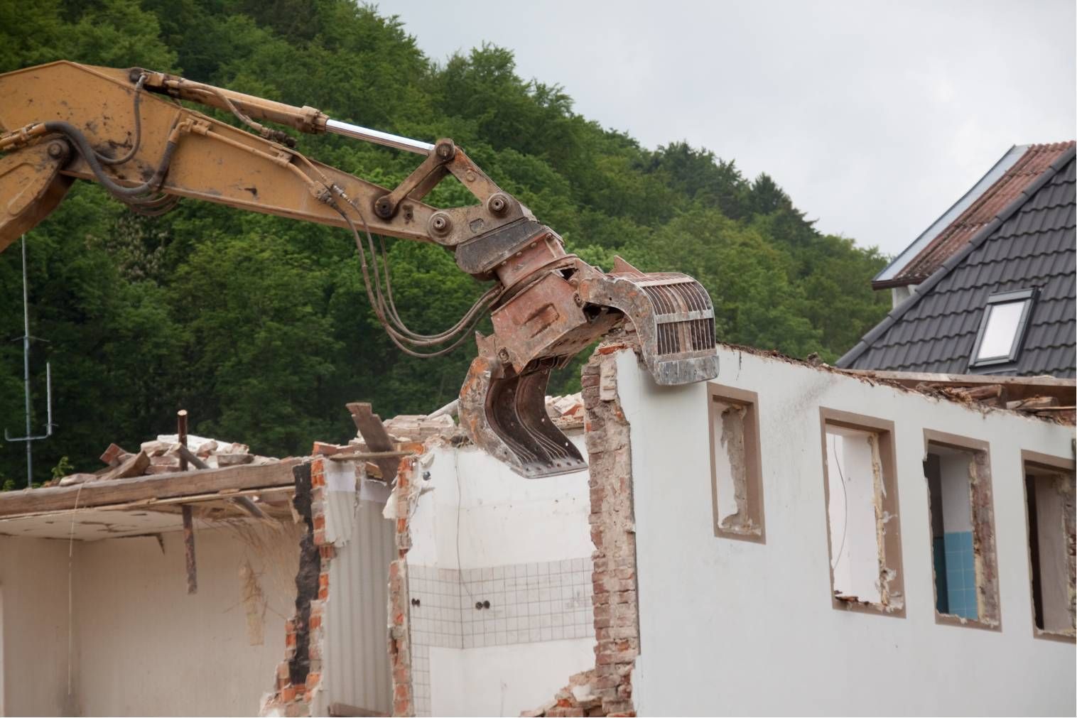Excavator demolishing a white house wall beside a roof and trees
