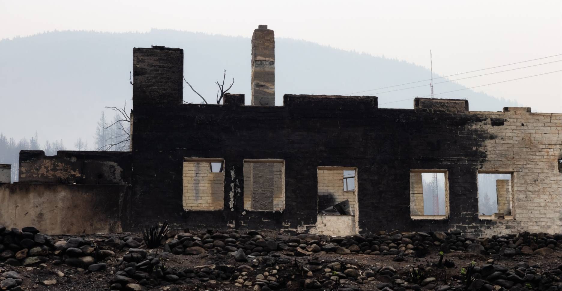 Burned-out house ruins with charred walls and collapsed roof against a smoky hillside background