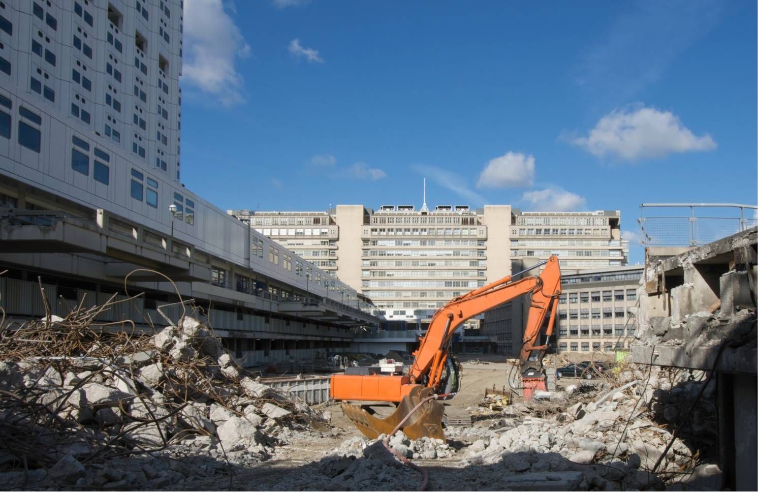 Orange excavator amid rubble at a demolition site between city buildings