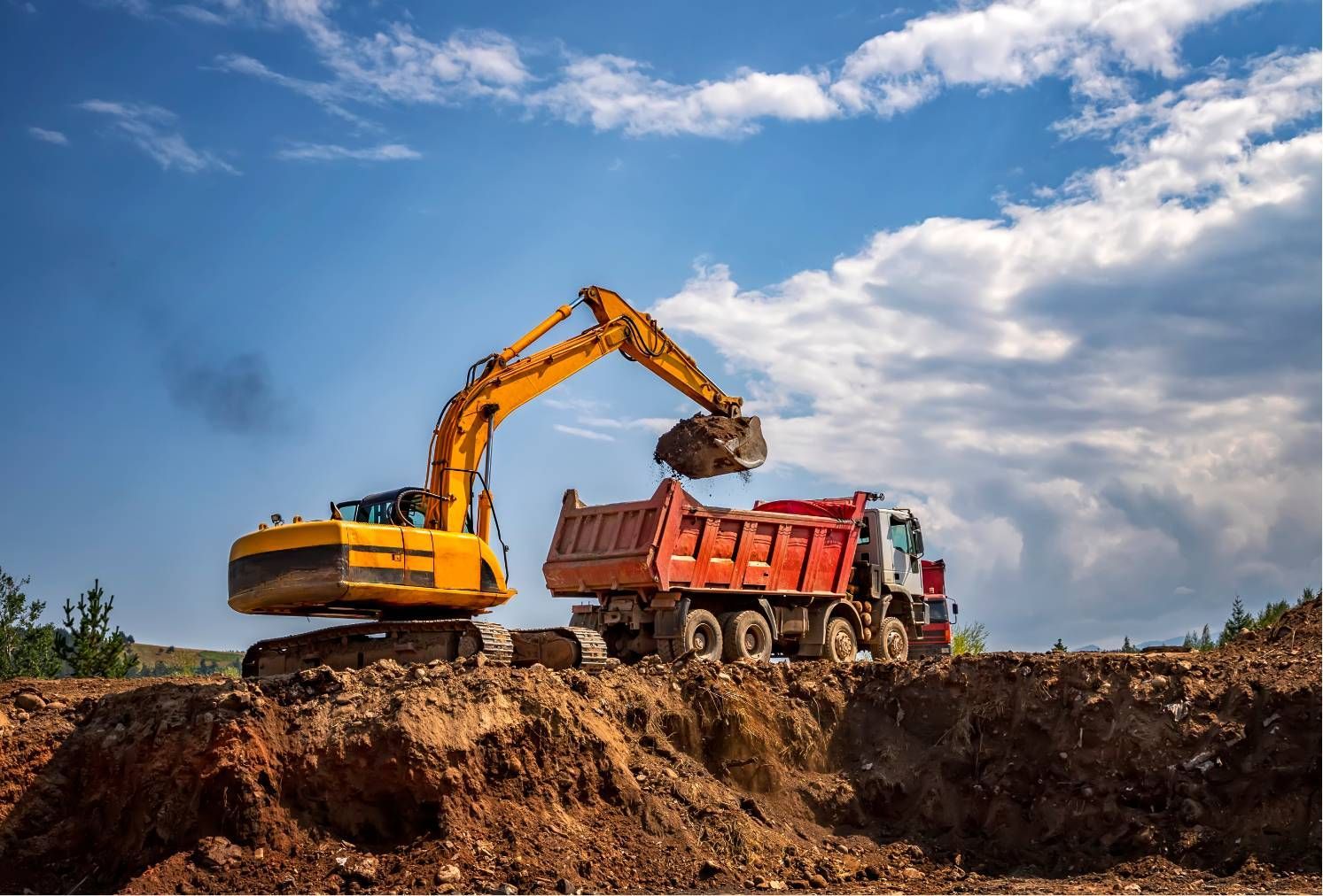 Yellow excavator loading dirt into a red dump truck at a construction site under a blue sky