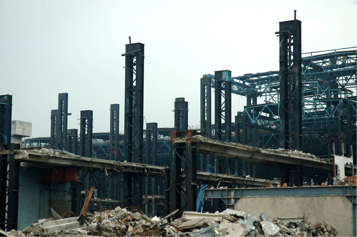 Ruins of a burnt industrial complex with twisted steel, collapsed structures, and debris under a gray sky