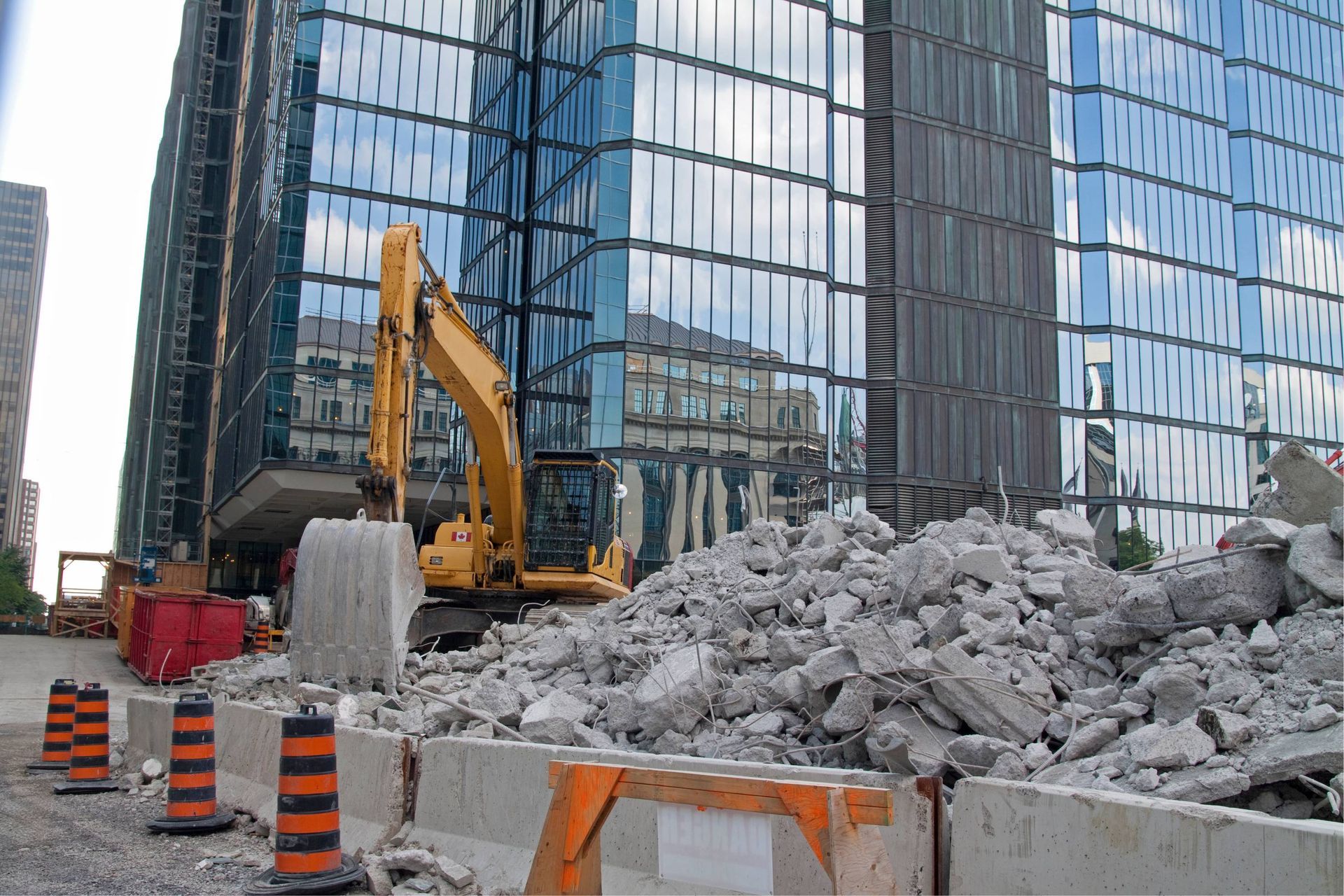 Construction site with excavator, rubble pile, traffic cones, and glass office buildings in the background