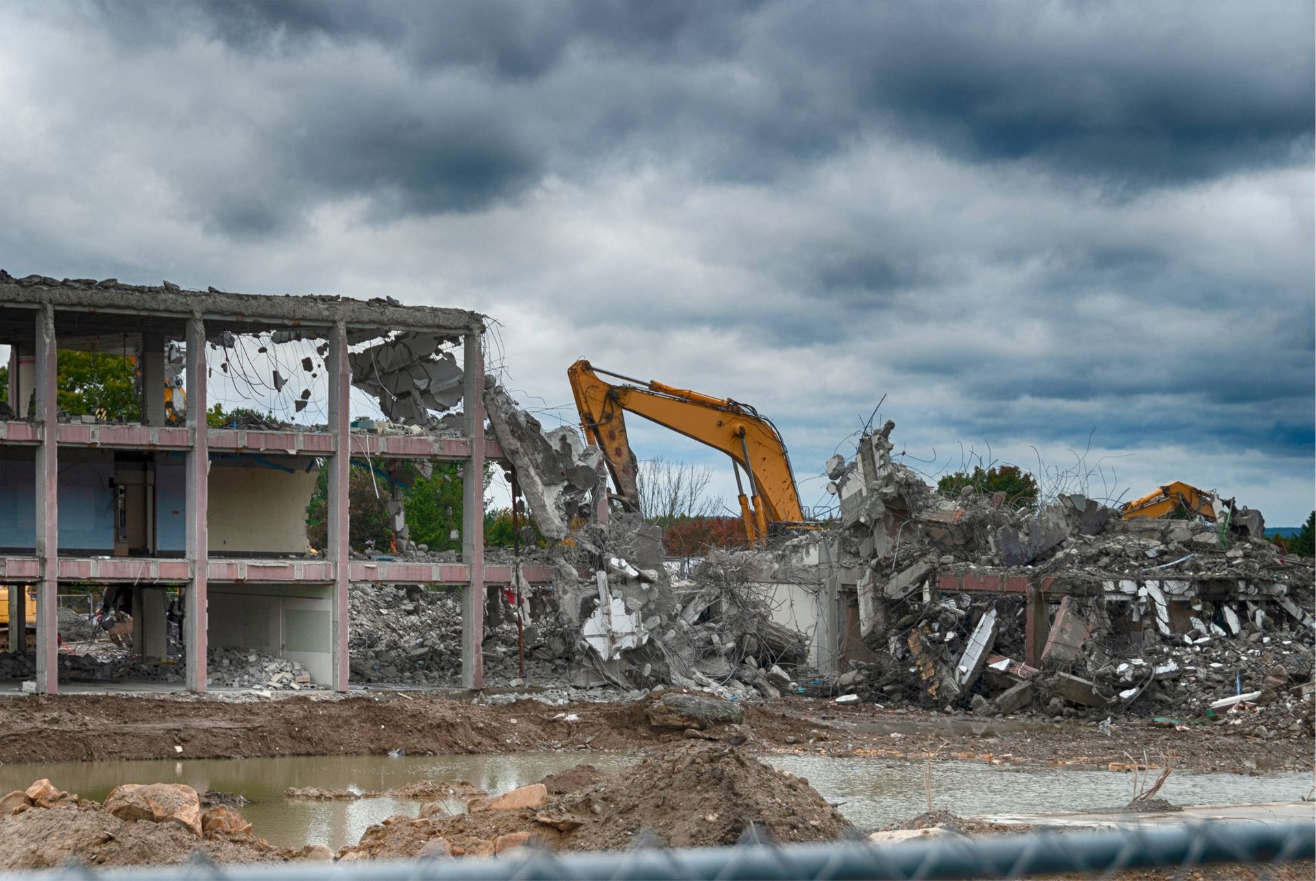 Demolition excavator tearing down a damaged building under dark storm clouds