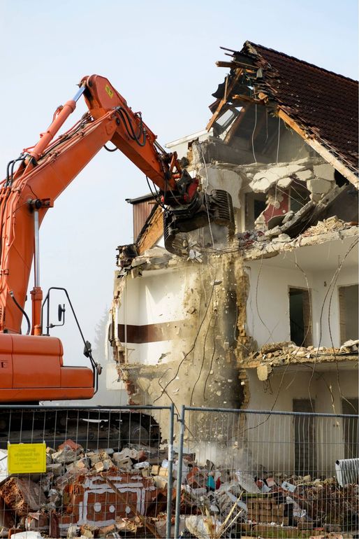Orange excavator demolishing a damaged house beside a fenced debris pile